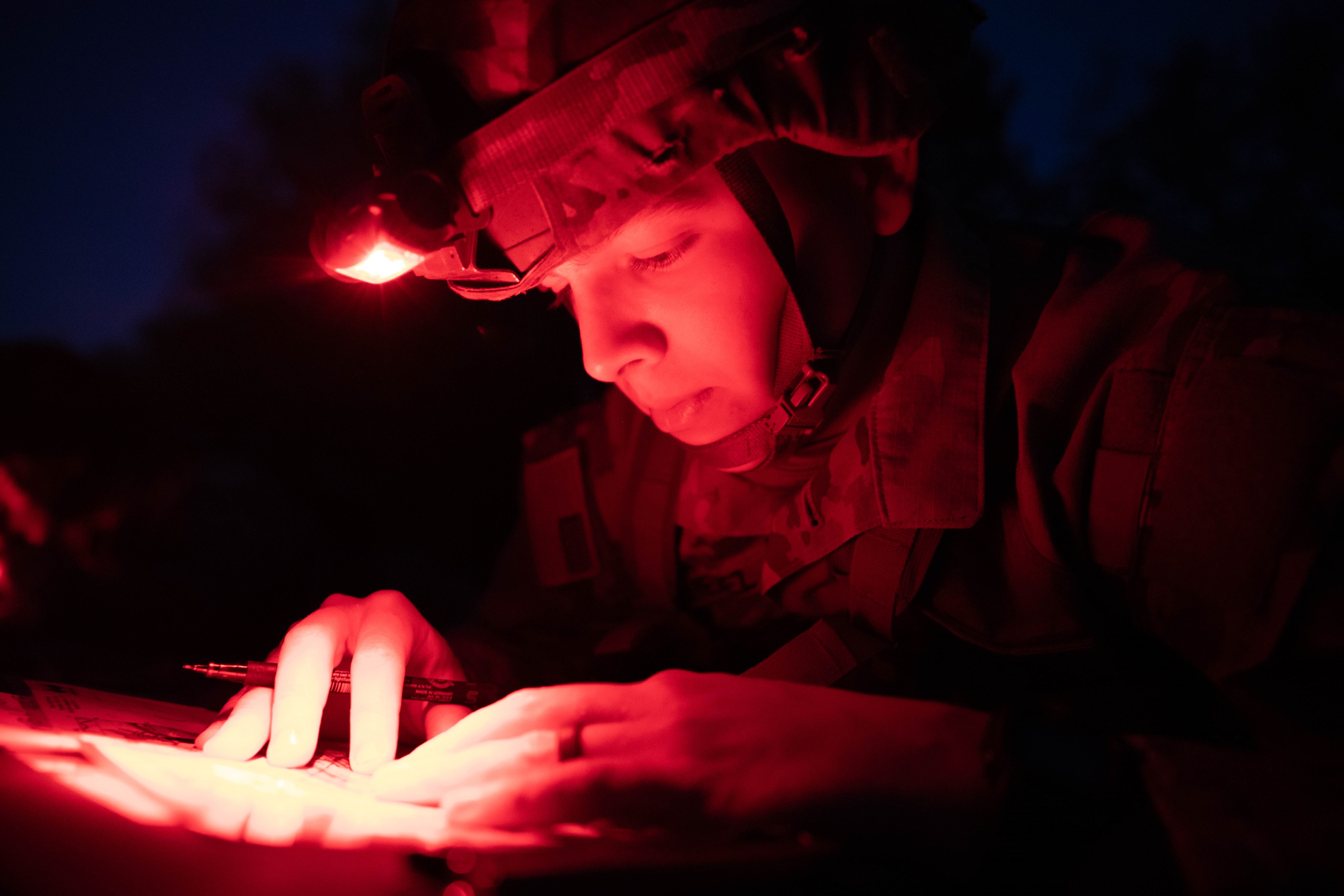 Under a reddish glow of light, a Soldier plots map points before being tested on night land navigation. 