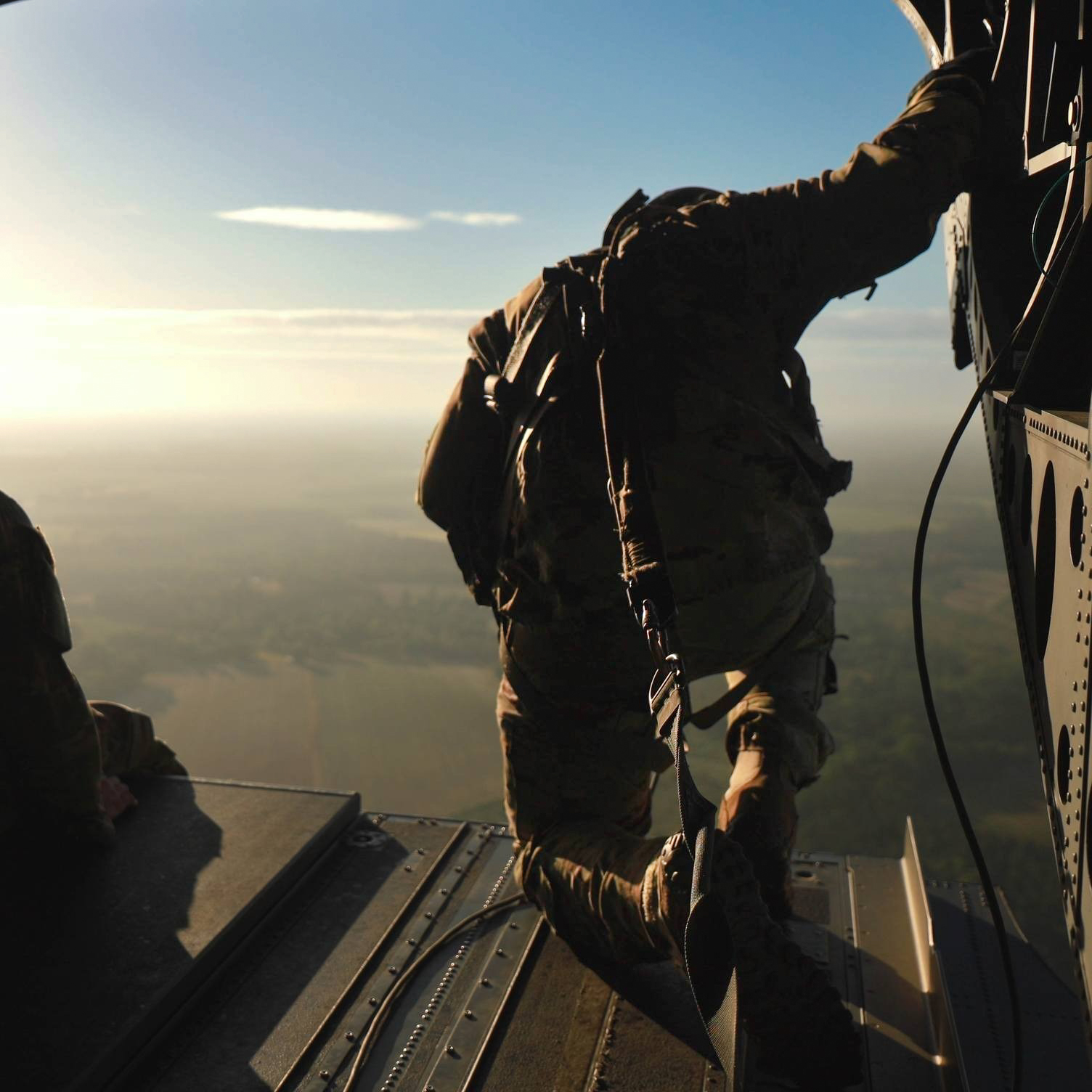 This photo shows a Soldier standing at the open rear ramp of a military aircraft, poised as if preparing to jump or observe the terrain below. The figure is silhouetted against a clear sky, and their gear is visible, emphasizing readiness and precision. 